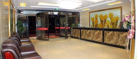 A hotel lobby with a reception desk, a waiting area with chairs, and a glass door entrance.