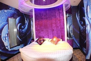 Ein Zimmer mit einem Bett, einem großen Spiegel und einer violetten Wand mit Struktur.