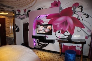Une chambre à coucher comprenant un lit, un téléviseur fixé au mur, une coiffeuse avec un miroir et un mur décoratif orné de motifs floraux.