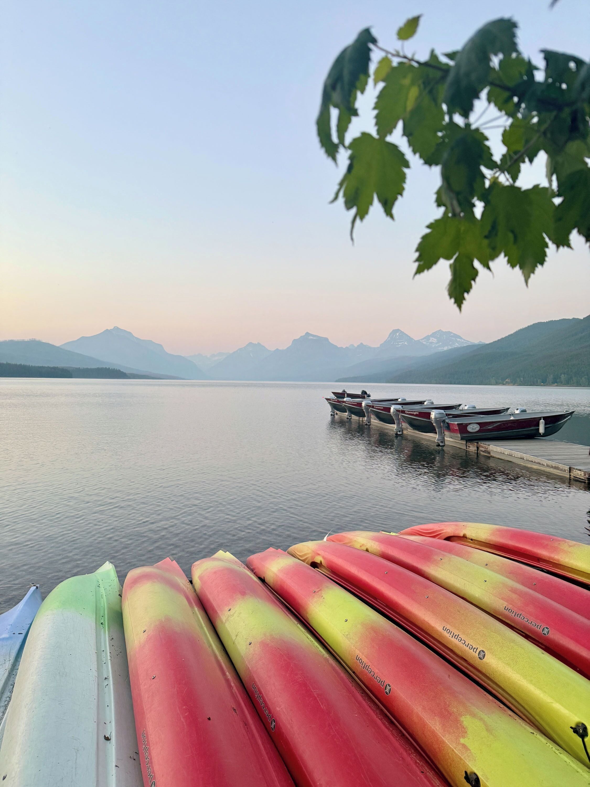 A row of colorful kayaks lined up at a dock with a calm lake and mountains in the background.