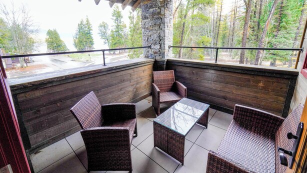 A covered patio with wicker furniture and a stone wall, overlooking a forested area. - Apgar Lookout Suites (West Glacier)