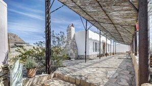 Une cour dallée de pierres, avec des marches menant à un bâtiment blanc, une tour de pierre et un espace ombragé agrémenté d’une pergola.