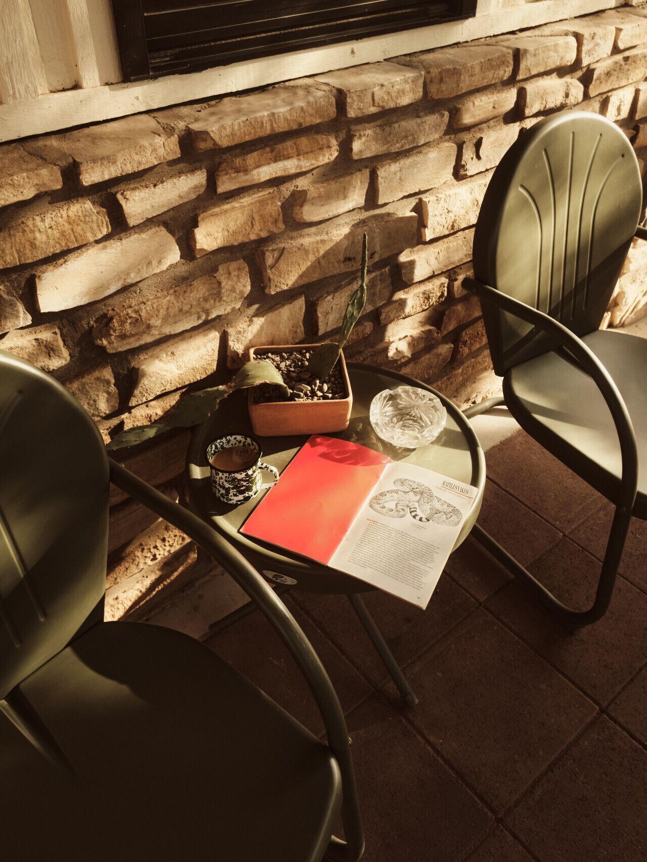 Un muro de piedra, dos sillas de metal, una mesa con un libro, una planta en maceta y una taza de café.