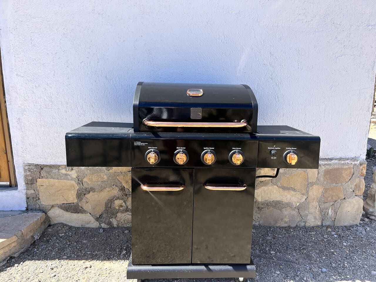 A black outdoor grill with four burners and a digital display, set against a stone wall and a white building.