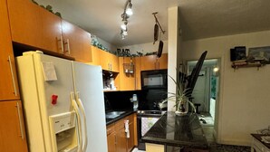 A compact kitchen with wooden cabinets, a white refrigerator, a black countertop, and a small dining area with a stool. - Waterfront Condo with Fishing, Paddleboard, and yard access. (North Miami)