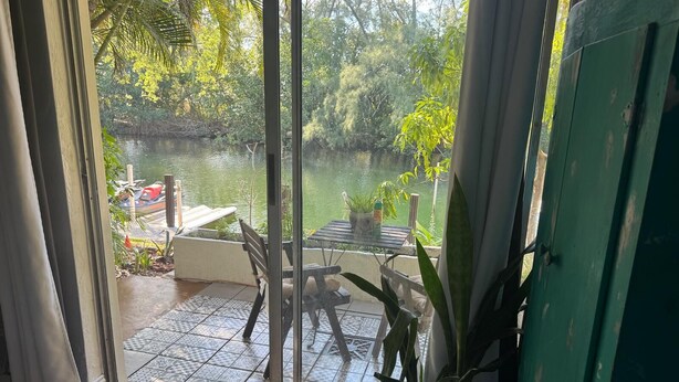 A room with a sliding glass door leading to a balcony with a view of a body of water, a table, chairs, and a potted plant. - Waterfront Condo with Fishing, Paddleboard, and yard access. (North Miami)