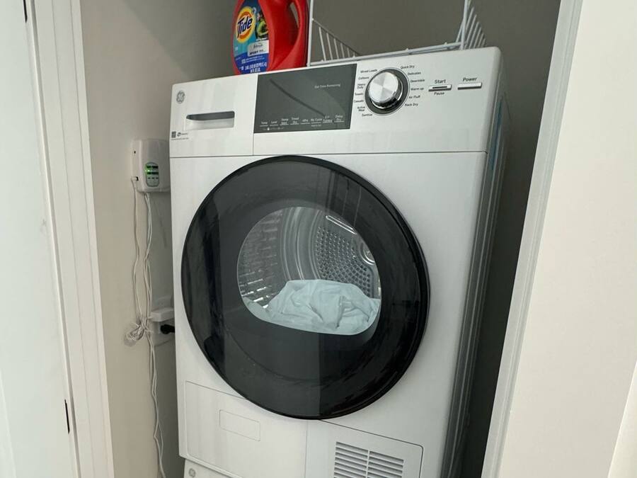 Una lavadora y secadora apiladas en un lavadero pequeño, con una puerta a la izquierda y un enchufe en la pared.