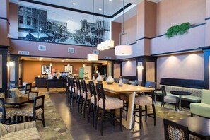 Een hotel lobby met een lange eettafel, diverse stoelen, een bar en een zithoek met banken en fauteuils.