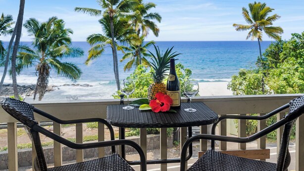 Un balcón con sillas de mimbre y una mesa, con vista a una playa con palmeras y el océano.