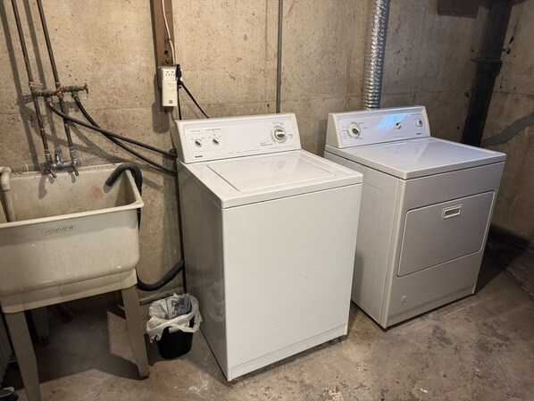 Dans une buanderie au sol en béton et aux tuyaux apparents, on trouve deux lave-linge à chargement frontal blancs, dotés de panneaux de commande, un évier utilitaire blanc et une poubelle noire.