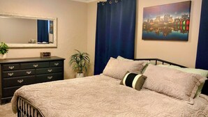 Un dormitorio con cama, cómoda, espejo, una pintura de paisaje urbano y cortina.