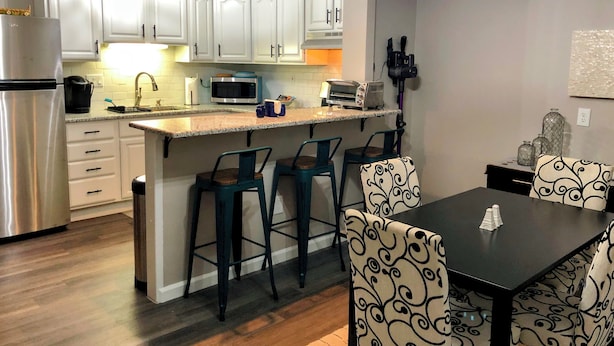 Una cocina moderna con armarios blancos, un refrigerador de acero inoxidable, un microondas y un tostador. Hay tres taburetes de bar y una mesa de comedor con sillas estampadas.