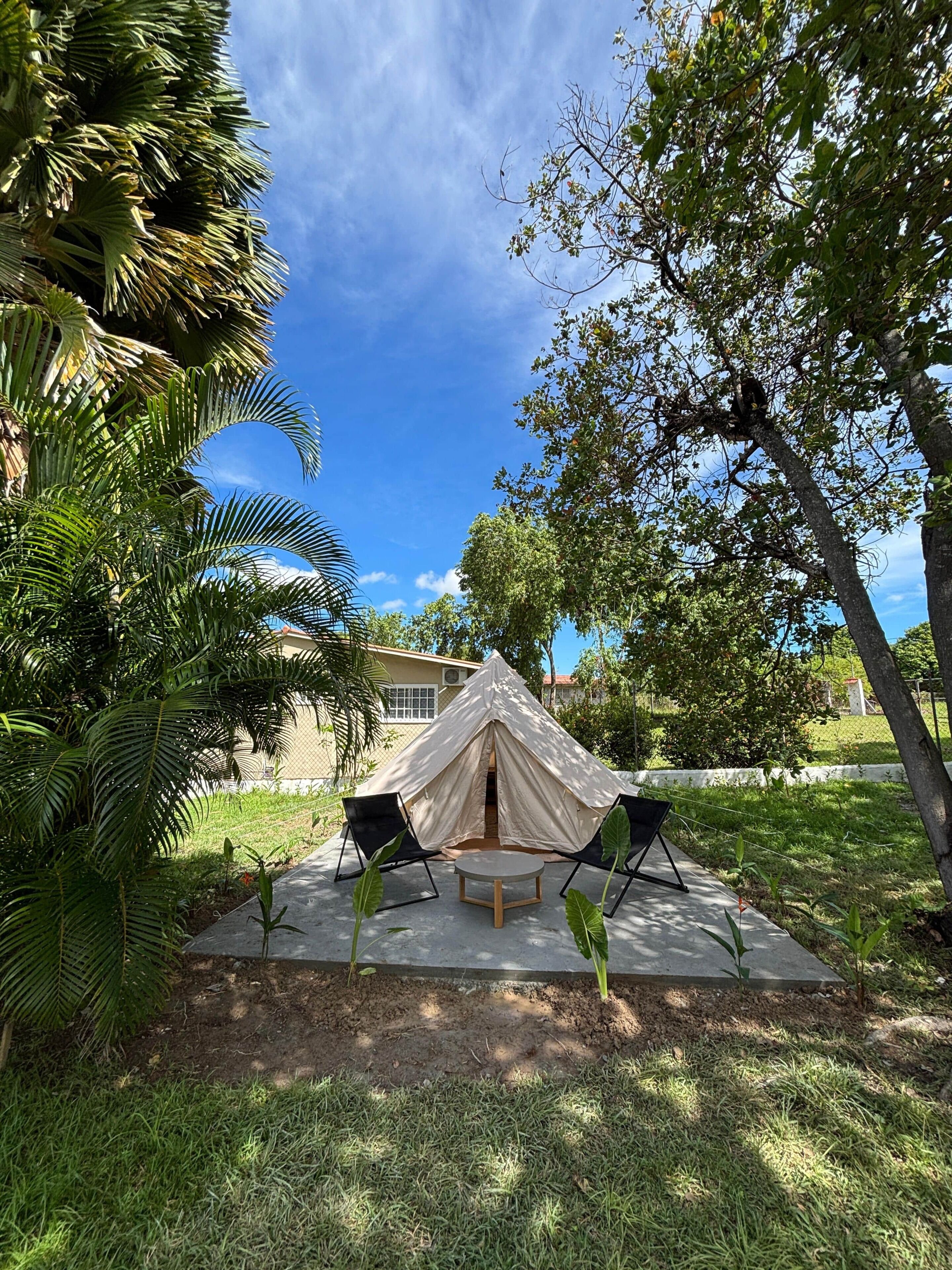 A tent with two chairs, surrounded by greenery and a building in the background.