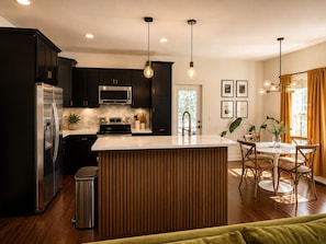Una cocina moderna con armarios oscuros, electrodomésticos de acero inoxidable, una isla central y una zona de comedor con sillas y mesa de madera.