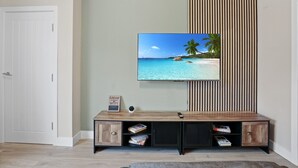 A living room with a flat-screen TV mounted on a wall, a wooden entertainment unit with books and a remote, and a door to the left.