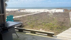 Een houten terras met een glazen tafel en stoelen, uitkijkend op een zandstrand en een heldere lucht.