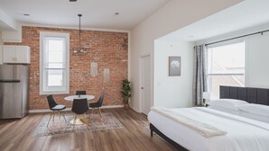 Een moderne slaapkamer met een groot bed, een eethoek met tafel en stoelen, een keuken met een koelkast, en een bakstenen muur.
