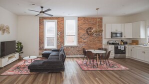 A modern living room with a sofa, a dining table, and a kitchen area with white cabinets and a microwave. - Hillsborough Castle- Efficiency Suite (Martinsburg)