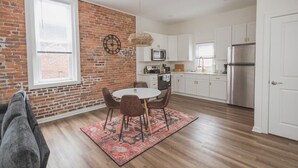 A kitchen with white cabinets, a brick wall, a round dining table with chairs, and a hanging light fixture. - Hillsborough Castle- Efficiency Suite (Martinsburg)