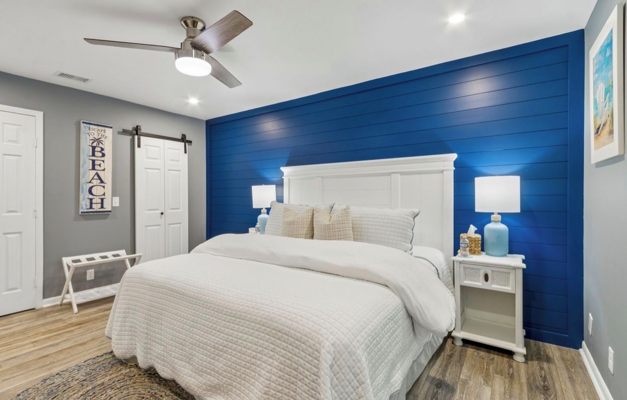 Une chambre avec un lit blanc, un ventilateur de plafond et un mur d’accent bleu.
