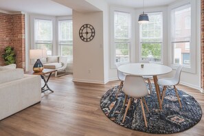 Un salon moderne avec un canapé blanc, une table ronde entourée de chaises, une horloge au mur et de grandes fenêtres équipées de stores.