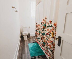Un baño estrecho con inodoro, un cubo de basura pequeño y un felpudo color turquesa.