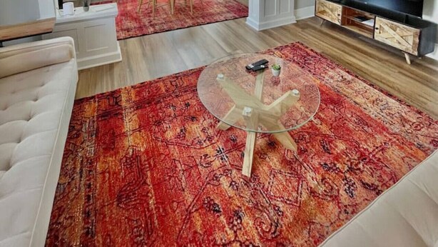 Un salon comprenant un téléviseur, une table basse en verre, un tapis à motifs rouges, un canapé blanc et un coin repas avec une table et des chaises blanches.