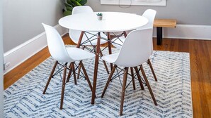 Un coin repas avec une table ronde, des chaises blanches, un luminaire suspendu en osier, une plante en pot et une fenêtre agrémentée de rideaux blancs.