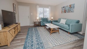 A living room with a TV, sofa, and coffee table, a patterned rug, and a window with blinds.