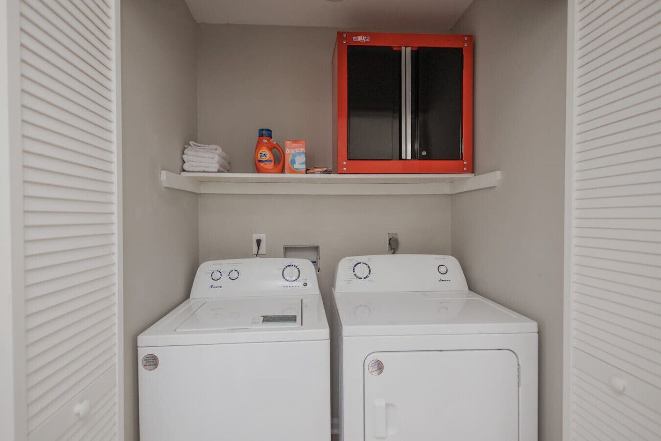 A laundry room with two white front-loading washing machines, a shelf with laundry detergent, and a red cabinet above.