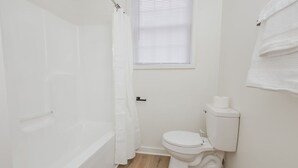 A small, white bathroom with a bathtub, toilet, and a window with blinds. - The Radiant Residence (Martinsburg)