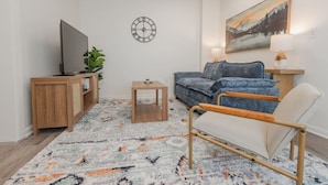 Ein Wohnzimmer mit Fernseher, Sofa, Sessel, kleinem Tisch und einer Wanduhr.