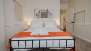 Una cama bien hecha con ropa de cama blanca y naranja, un tapiz de macramé y tres cuadros enmarcados sobre la cama.