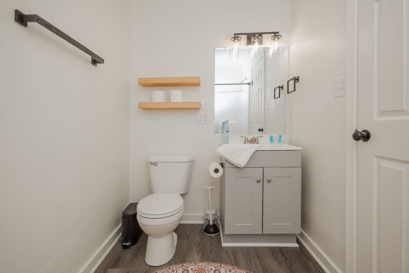 A small bathroom with a toilet, sink, and mirror. There are wooden shelves above the toilet and a black trash bin next to the toilet.