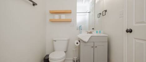 A small bathroom with a toilet, sink, and mirror. There are wooden shelves above the toilet and a black trash bin next to the toilet.