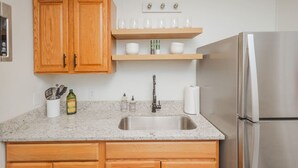 Una cocina con armarios de madera, un lavamanos de acero inoxidable, un refrigerador y una estantería con vasos y cuencos.