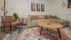 Una sala con un sillón de cuero marrón, un sofá beige, una mesita redonda de madera y un tapete estampado.