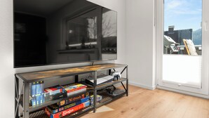 Un saló modern amb un gran televisor de pantalla plana fixat a la paret, una taula de centre de fusta i una biblioteca plena de jocs de taula.