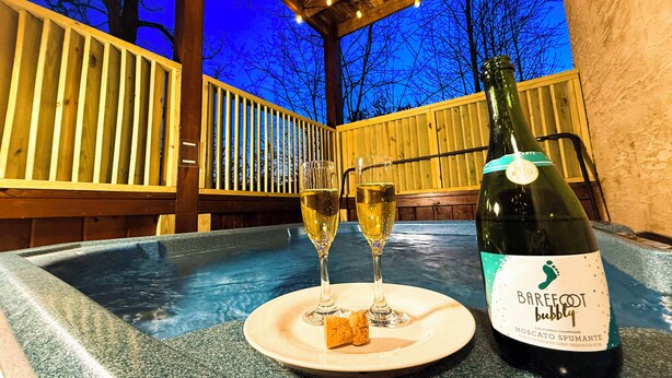 Una terraza de madera con un jacuzzi, dos copas de champán y una botella de Barefoot espumoso sobre una mesa.