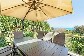 Eine Terrasse mit Tisch und Stühlen unter einem großen Sonnenschirm, mit Blick auf eine Küstenlandschaft.