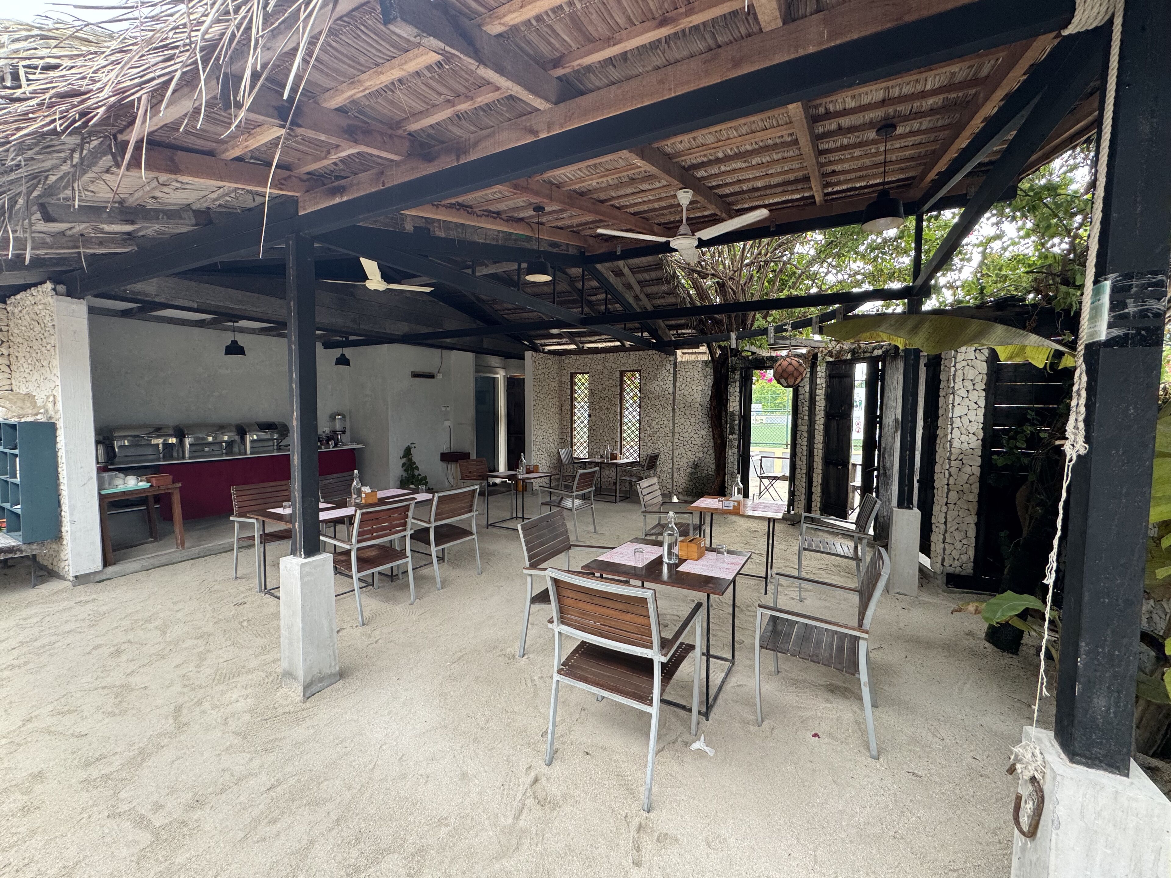 An outdoor dining area with tables and chairs, a ceiling fan, and a kitchen area in the background.