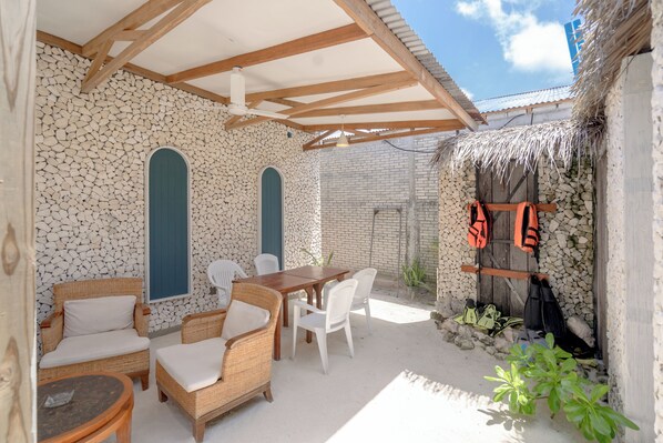 A covered outdoor seating area with wicker furniture and a table, a stone wall with a blue door, and a thatched roof structure with life jackets and shoes.