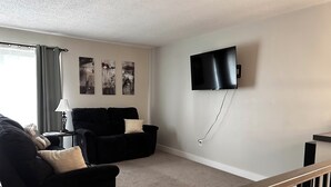 A living room with a black sofa, a small table with a lamp, and a flat-screen TV mounted on the wall.