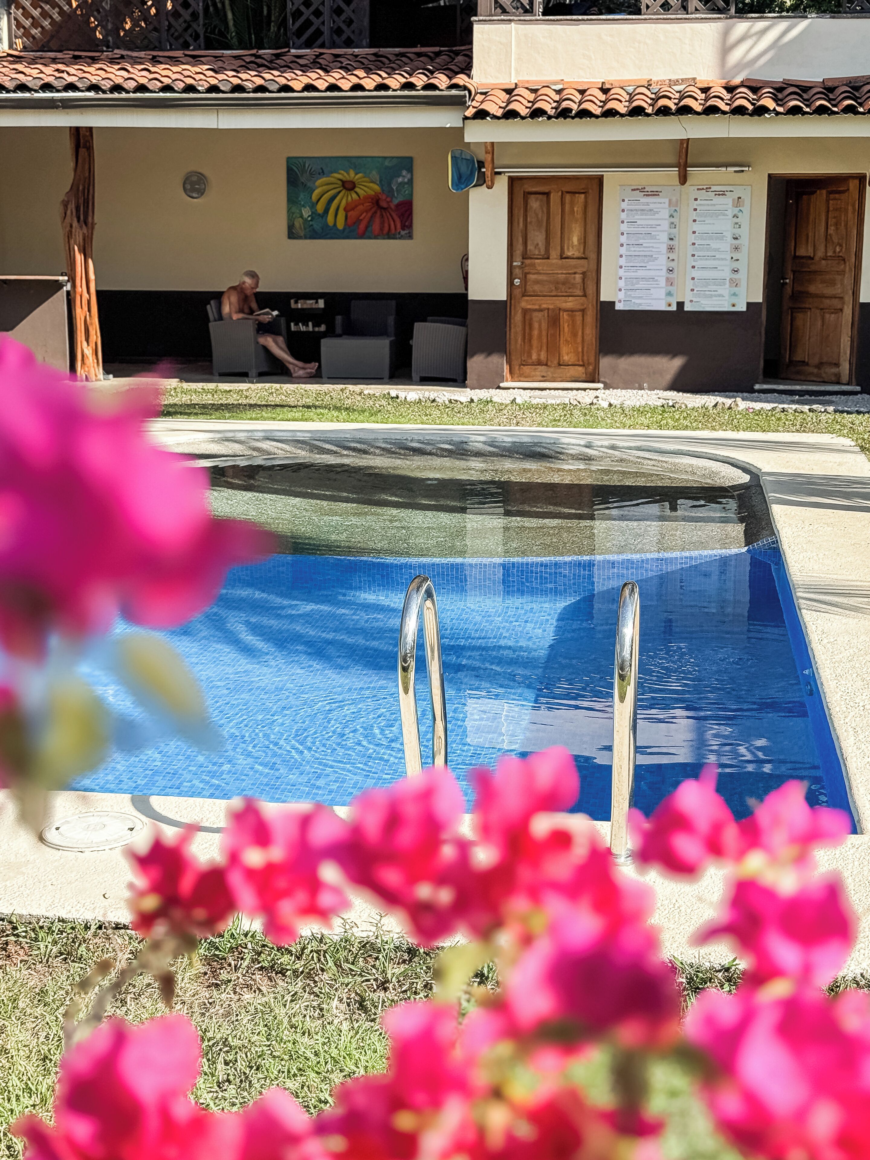Una piscina con scalette in metallo, circondata da un'area verde e un edificio con tetto in tegole e un murale.