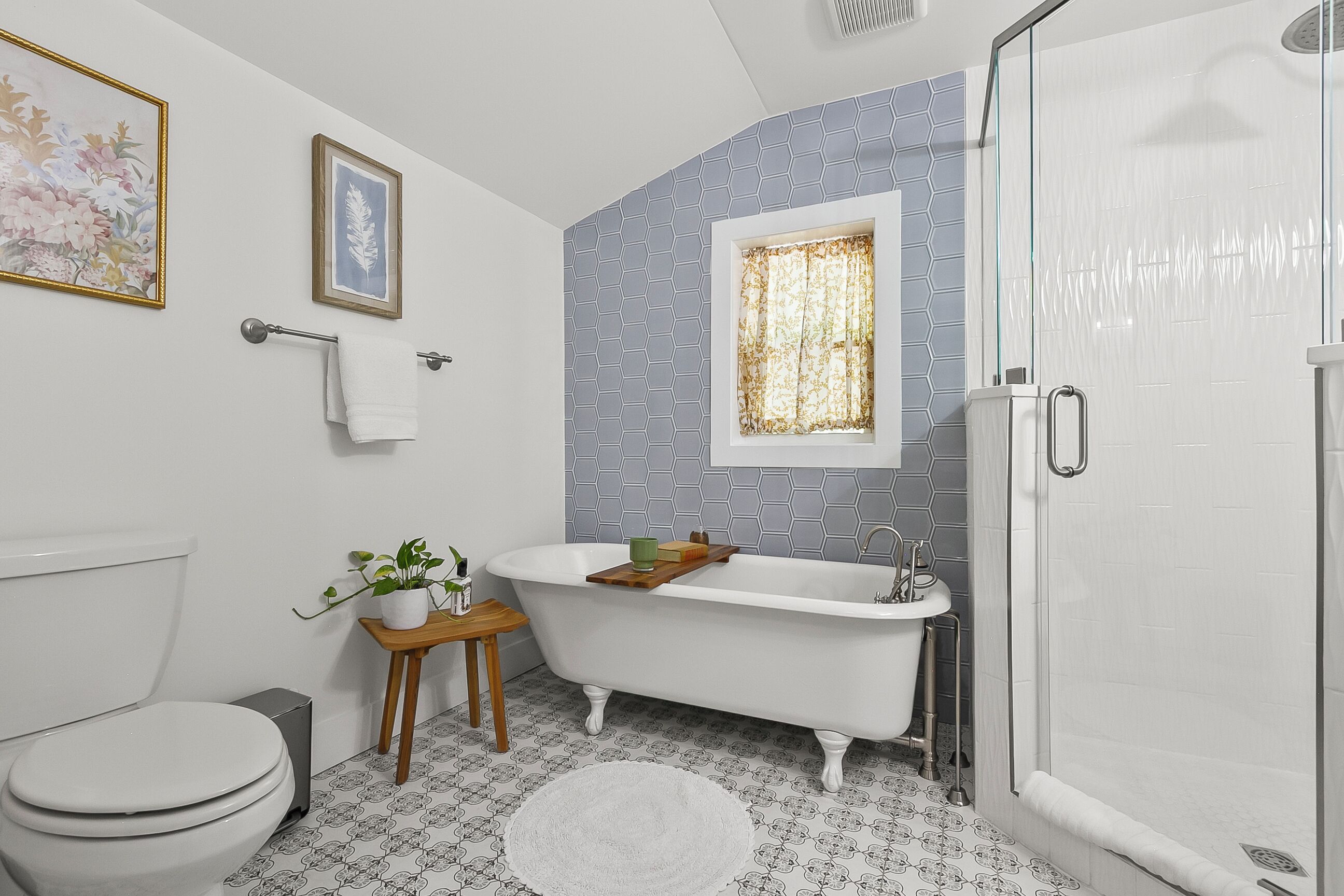Un baño con una bañera blanca, un inodoro, un taburete pequeño de madera con una planta y una ventana con cortinas estampadas.