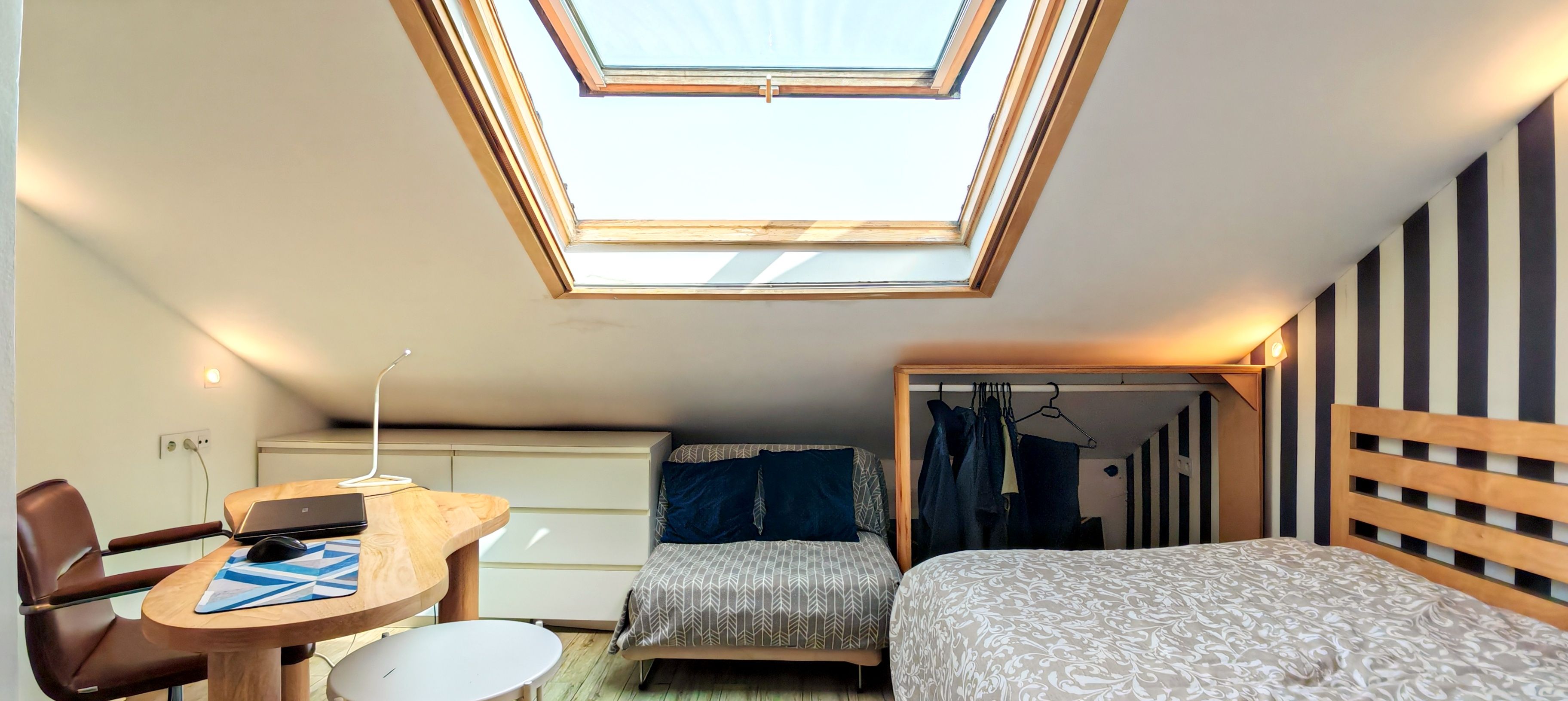 Una habitación compacta con tragaluz, un escritorio con una computadora portátil, una cama con ropa de cama estampada y un armario con paneles a rayas.