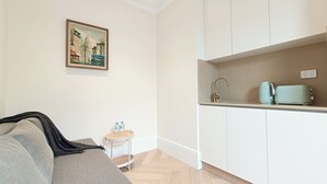 Een compact woongedeelte met een kitchenette, een klein tafeltje met waterflesjes en een schilderij aan de muur.