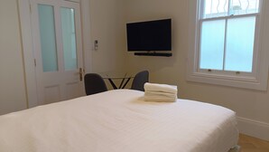 Een slaapkamer met een bed, een aan de muur bevestigde televisie, een raam met zonwering en een deur met geëtste glaspanelen.