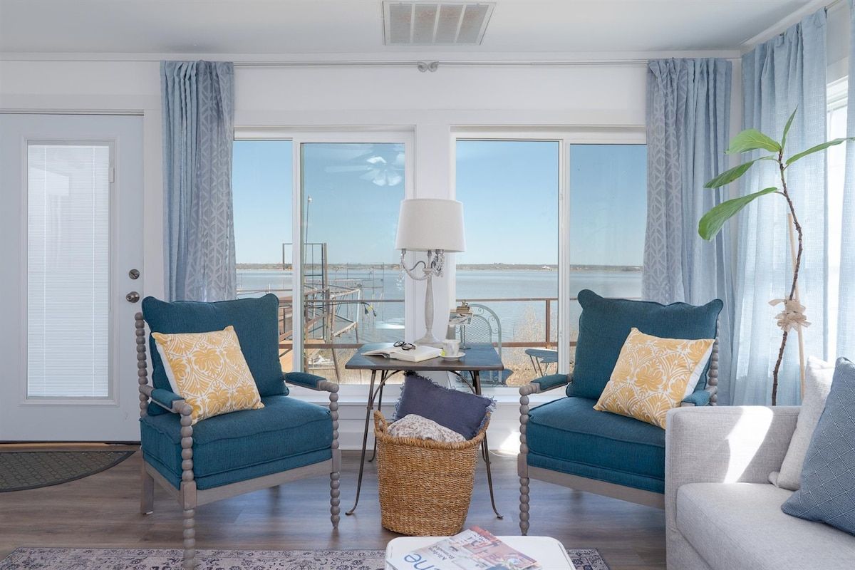 Een kamer met twee blauwe fauteuils, een klein tafeltje met een lamp, en uitzicht op zee door grote ramen.