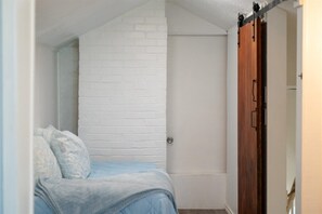 Une chambre avec un mur en briques blanches, une porte en bois et un lit recouvert d’une couverture bleue.